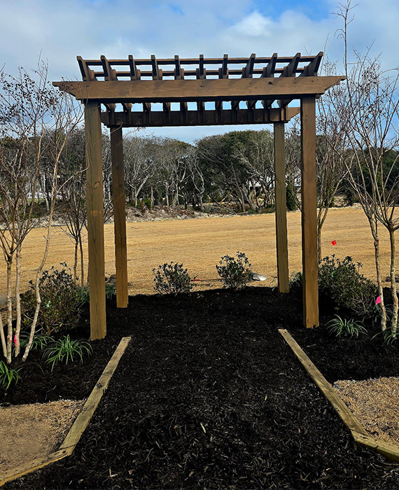 Custom wooden pergola installed with mulched planting beds, ornamental shrubs, and a defined garden pathway to enhance an outdoor gathering space.