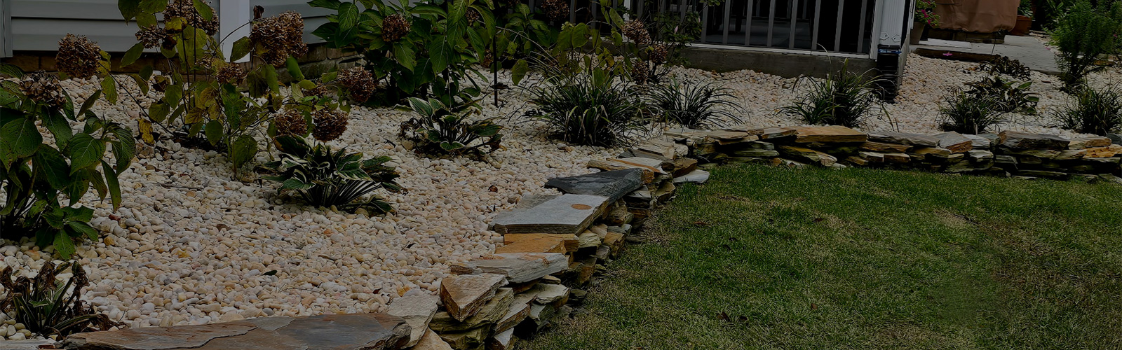 Residential landscape with stacked natural stone edging, decorative river rock, perennial plantings, and neatly maintained garden beds.